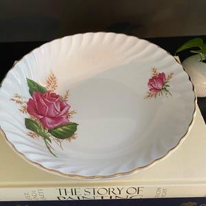 Peut inclure: Bol en porcelaine blanche avec un bord festonné et une bordure dorée. Le bol est décoré de roses roses et de feuilles vertes. Le bol est posé sur un livre.