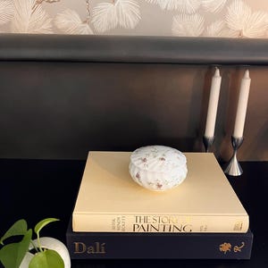 Peut inclure: Une petite boîte à bijoux ronde à motifs floraux avec un couvercle est posée sur une pile de livres. Les livres sont intitulés "The Story of Painting" et "Dali". Deux bougies blanches dans des bougeoirs argentés sont en arrière-plan.