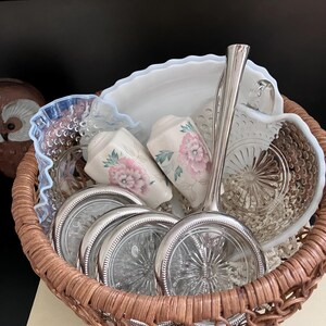 May include: A woven basket filled with vintage items. Includes a white and pink floral-patterned salt and pepper shaker set, glass coasters with silver rims, a glass serving dish, and a silver serving spoon. The basket is brown and contains a variety of antique items.