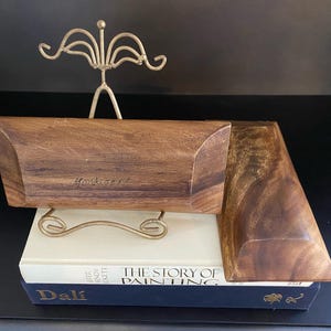 Puede incluir: Una caja de madera con tapa, apoyada sobre una pila de libros. La caja es de color marrón cálido y tiene una forma geométrica única. El texto "Monkwood" es visible en la caja. Un soporte decorativo de metal dorado está detrás de la caja.