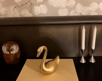 Può includere: Una statuetta di cigno dorato è appoggiata su una pila di libri, tra cui "The Story of Painting". Sono visibili anche una statuetta di gufo e due candelabri argentati con candele bianche. Lo sfondo presenta una carta da parati a motivi.