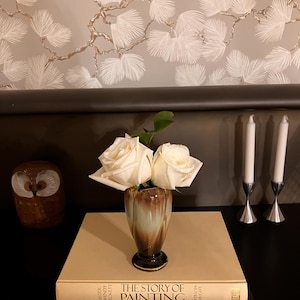 Pode incluir: Um pequeno vaso castanho e verde contém duas rosas brancas. O vaso está em cima de um livro bege intitulado "THE STORY OF PAINTING" e um livro preto com "Dali" em letras douradas. Uma figura de coruja de madeira e dois castiçais prateados também são visíveis.