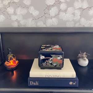 May include: A black lacquered square box with a glossy finish, decorated with intricate floral patterns in red, white, and green. The box sits atop two stacked books, one titled The Story of Painting and the other labelled with the name Dalí.