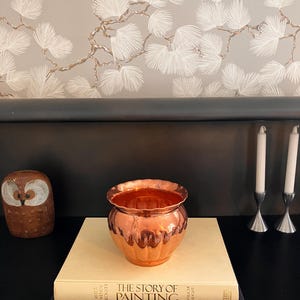 May include: A copper-colored, scalloped-edge vase sits atop a stack of books, with an owl figurine to the left and two silver candle holders to the right. The background features a gray wall with a white and gold leaf pattern.