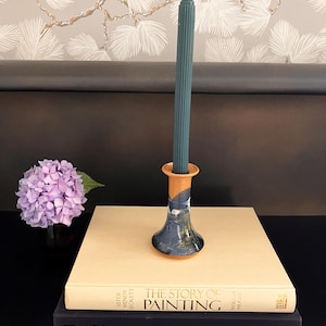 May include: A teal candlestick with a fluted design holds a teal candle. The candlestick has a blue and tan painted design. It sits atop two books, one titled "The Story of Painting". A purple hydrangea is in a vase nearby.