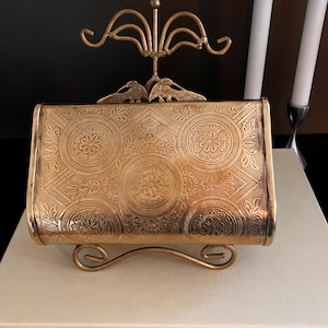 Könnte beinhalten: Eine goldfarbene, rechteckige antike Clutch mit einem verzierten Blumenmuster. Die Clutch wird auf einem dekorativen goldenen Ständer ausgestellt. Ein kleiner, dekorativer goldener Vogel befindet sich oben auf der Clutch. Zwei weiße Kerzen sind im Hintergrund.