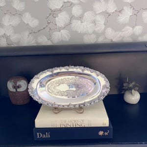 Peut inclure: Un plateau de service ovale en argent avec un motif floral gaufré orné au centre et un bord festonné décoratif présentant des détails de feuilles complexes. Le plateau est présenté sur un support posé sur deux livres reliés.