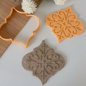 May include: Three cookie cutters and a cookie. The cutters are orange and shaped like a decorative tile. One cutter is a frame, another is a solid tile shape, and the third is a cookie with an embossed design. The cookie is light brown.