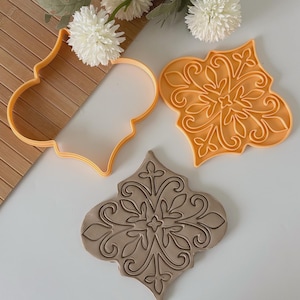 May include: An orange cookie cutter and a pressed cookie with an ornate, floral design. The cookie cutter is in the shape of a quatrefoil. The pressed cookie is tan and has a detailed pattern. White flowers are in the background.