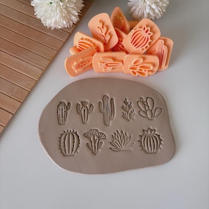 May include: A collection of peach-colored cactus-shaped stamps and a clay slab with impressed cactus designs. The stamps are arranged on a white surface, with a wooden board and white flowers in the background. The clay slab features various cactus and succulent impressions.