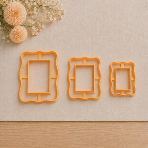 May include: Three orange plastic cookie cutters in descending sizes, each featuring a rectangular center surrounded by a decorative scalloped frame border. The cutters are arranged horizontally, showcasing a consistent design with structural support bars.