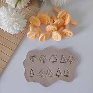 Può includere: Una collezione di timbri a forma di albero arancione chiaro con manici, insieme a una lastra di argilla stampata con vari disegni di alberi. I timbri sono per l'artigianato o la decorazione, con un focus su elementi a tema natura.
