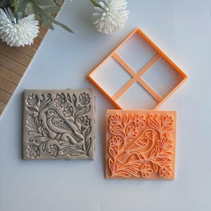 May include: Square clay stamp with a bird and floral design. The stamp is gray and has a detailed relief pattern. An orange square frame and a matching orange stamp with the same design are also present.