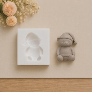Op de afbeelding: Een witte vierkante siliconen mal met een uitsparing in de vorm van een teddybeer met een slaapmuts, naast een afgewerkt grijs teddybeerfiguurtje met een bijpassende muts en trui, met kleine lichtroze bloemetjes in de hoek.