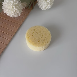 Op de afbeelding: Een ronde, lichtgele spons met een poreuze textuur. De spons is cilindrisch en ligt op een wit oppervlak. Decoratieve witte bloemen en een houten oppervlak zijn op de achtergrond.