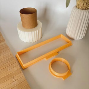 May include: An overhead shot shows a collection of craft supplies. There's a cylindrical cardboard tube, a white, ribbed, circular base, and two orange plastic cookie cutters. A white vase with wheat stalks is also visible.