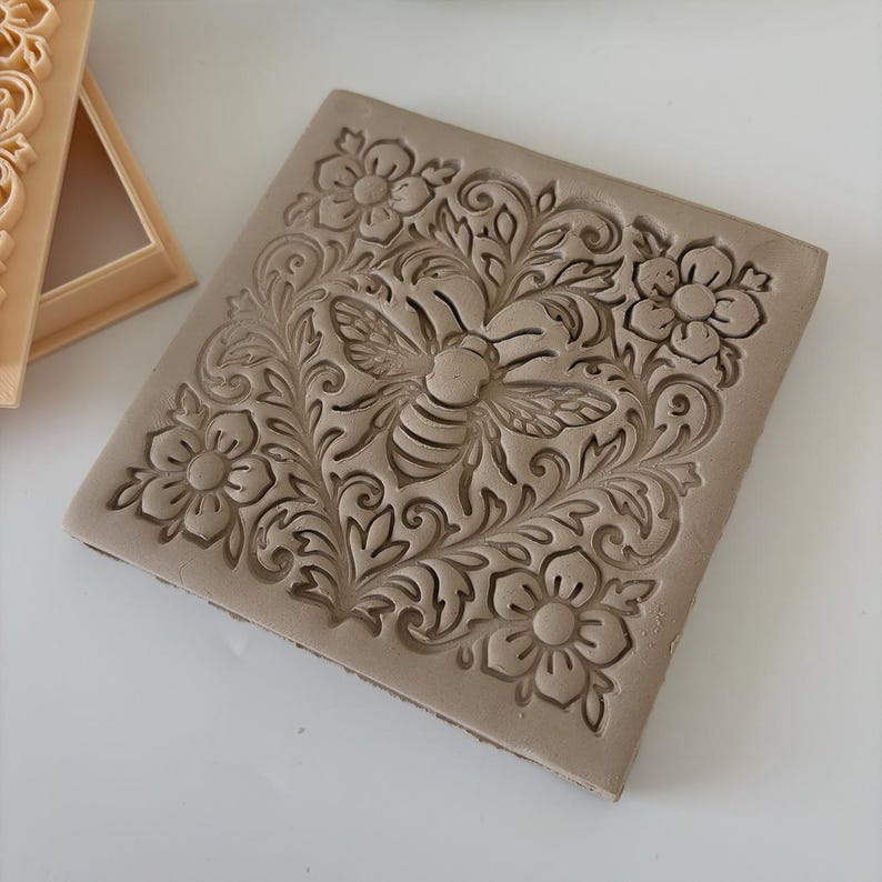 May include: A square, beige stamp with a detailed bee and floral design. The bee is centered, surrounded by flowers and leaves. The stamp is likely for crafts or baking, adding a decorative element to projects.