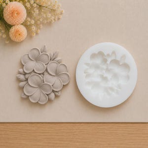 Op de afbeelding: Een ronde witte siliconen mal met een bloemmotief naast een grijze kleiafgietsel van hetzelfde ontwerp. Het afgietsel toont een cluster van drie opvallende vijfbladige bloemen omringd door kleinere knoppen en bladeren in reliëf.