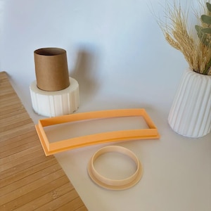 May include: An arrangement of baking tools and decorative elements. A light orange rectangular cookie cutter and a round cookie cutter are in the foreground. A brown cardboard tube sits atop a white, fluted base. A white vase holds wheat stalks.