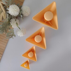 May include: Four orange-yellow triangular containers of varying sizes, each with a circular, light-yellow center piece. The containers are arranged diagonally on a white surface, with decorative white flowers and greenery in the background.