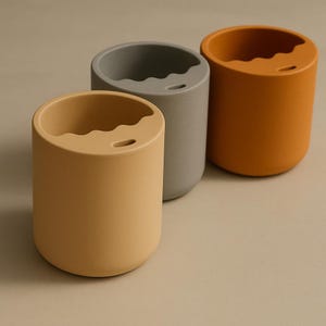 Puede incluir: Tres recipientes cil&iacute;ndricos en tonos amarillo, gris y naranja. Cada uno tiene un borde superior ondulado y un peque&ntilde;o agujero cerca del borde. Los recipientes est&aacute;n dispuestos en fila, mostrando su dise&ntilde;o simple y moderno.