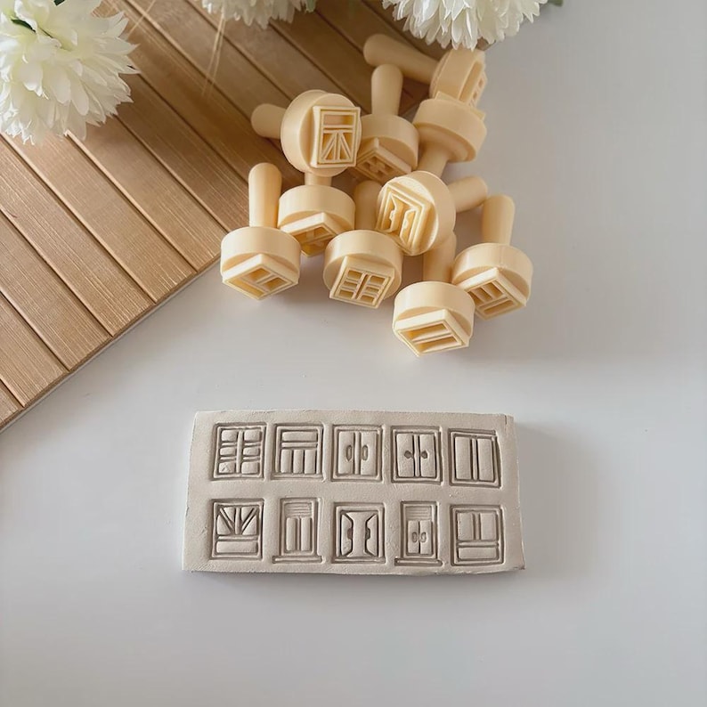 Peut inclure: Un ensemble de tampons beiges en forme de fen&ecirc;tre avec des poign&eacute;es, ainsi qu'un moule rectangulaire en argile blanc cass&eacute; pr&eacute;sentant divers motifs de fen&ecirc;tres. Les tampons sont &eacute;parpill&eacute;s, tandis que le moule affiche des contours de fen&ecirc;tres d&eacute;taill&eacute;s.