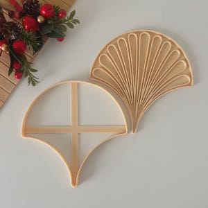 May include: Two beige plastic cookie cutters in a fan shape. One cutter has a solid fan design, and the other has a cross-shaped design. The cutters are on a white surface. The image also includes a decorative holiday arrangement.