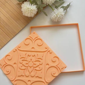 May include: An orange, square-shaped silicone mold with a floral and scroll design. The mold is next to a square frame and a wooden surface. White flowers are in the background.