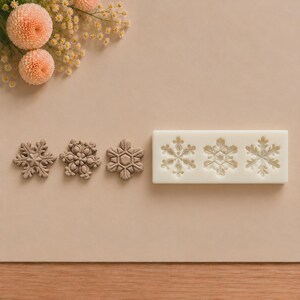 May include: A rectangular white silicone mold featuring three distinct snowflake cavities, accompanied by three individual light brown snowflake-shaped clay or resin castings displayed to the left of the mold on a neutral surface.