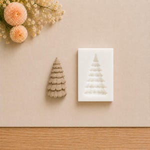 May include: A tan-colored, cone-shaped tree figurine with layered, scalloped branches next to a rectangular white silicone mold featuring a recessed, matching tree-shaped cavity. The items are arranged on a plain, light-colored surface.