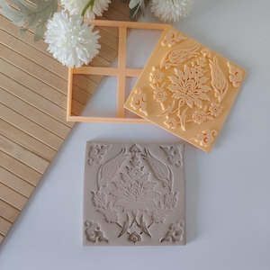 May include: An orange square cookie cutter and a beige square cookie stamp with a floral design. A beige square cookie with the same floral design is also present. The items are on a white surface with a bamboo mat and white flowers.