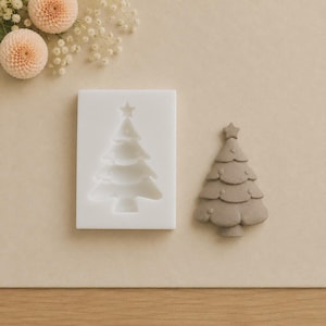 Könnte beinhalten: Eine rechteckige weiße Silikonform mit einer vertieften Weihnachtsbaumform mit Sternspitze, neben einem fertigen, massiven grauen, dreistufigen Weihnachtsbaum-Ornament mit einem Stern an der Spitze und abgerundeten Astkanten.
