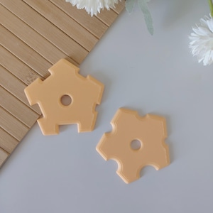 May include: Two light orange, five-sided coasters with a central hole and notched edges. The coasters are on a light gray surface, with a bamboo mat and white flowers in the background. The coasters have a geometric design.