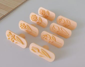 May include: A collection of eight light orange flower-shaped cookie stamps. The stamps feature various floral designs, including roses, tulips, and daffodils. Each stamp has a small handle on the back for easy use. The stamps are arranged on a light gray surface.