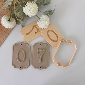 May include: A collection of beige and tan number-shaped cookie cutters. The set includes the numbers 0 and 7, along with a blank tag-shaped cutter. The cutters are made of plastic and are suitable for baking or crafting.