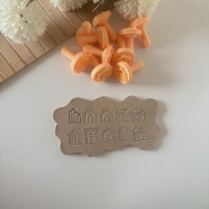 May include: A selection of orange plastic house-shaped stamps and a clay slab with house imprints. The stamps are scattered on a white surface, with a wooden tray and white flowers in the background. The clay slab has multiple house designs.