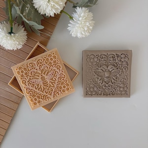 Könnte beinhalten: Zwei quadratische, dekorative Seifenformen. Eine ist hellbraun mit einem Bienen- und Blumendesign. Die andere ist grau mit dem gleichen Design. Die Formen sind von weißen Blumen und einer Holzoberfläche umgeben.