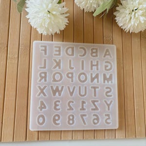 May include: A clear silicone mold with the alphabet and numbers. The mold is rectangular and features the letters A through Z, along with the numbers 0 through 9. The letters and numbers are in a sans-serif font. The mold is on a light brown wooden surface.