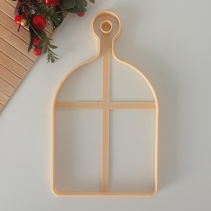 May include: A light beige cookie cutter in the shape of a serving board with a handle and a cross design. The cutter is made of plastic and is on a white surface. A decorative sprig of red berries and green leaves is in the upper left corner.