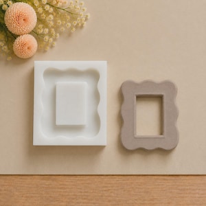 May include: A white rectangular silicone mold with a scalloped edge design and a central rectangular cavity, positioned next to a finished light brown rectangular frame with matching scalloped edges, both featuring a smooth, matte texture.