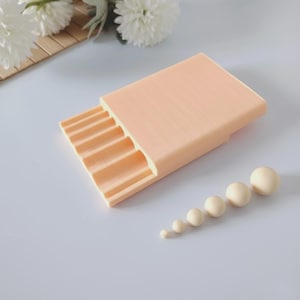 May include: A peach-colored, rectangular box with a series of curved slots on one side, and a row of cream-colored spheres of varying sizes. The box has a textured surface. The spheres are arranged in a diagonal line.