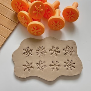 Op de afbeelding: Oranje sneeuwvlokvormige stempels en afdrukken in klei. De stempels hebben een handvat en een sneeuwvlokontwerp aan de onderkant. De klei heeft acht sneeuwvlokafdrukken. De stempels en klei liggen op een witte ondergrond.