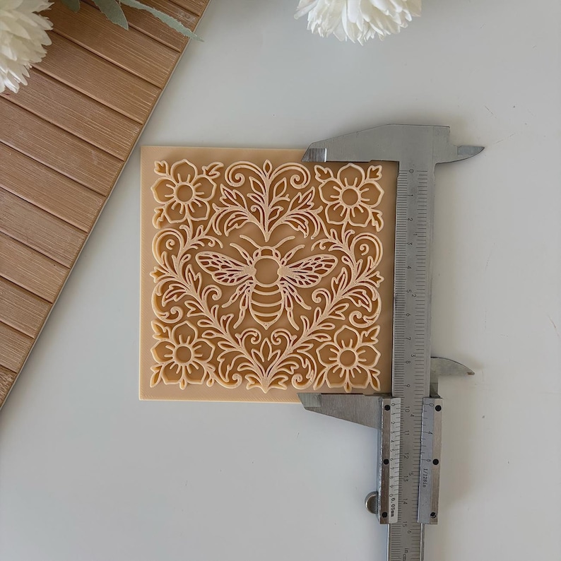 May include: A square, tan-colored craft item with a detailed bee and floral design. The design is cut out, creating a relief effect. A metal caliper is positioned next to the item, showing measurements.