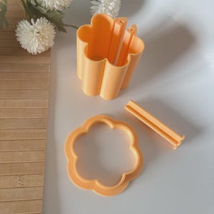 Puede incluir: Un juego de cortadores de galletas en forma de flor de color naranja. El juego incluye un cortador en forma de flor, un recipiente en forma de flor y dos piezas rectangulares. Los artículos son de plástico y están sobre una superficie blanca.