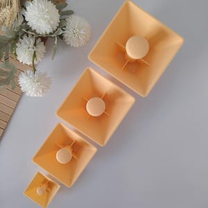 Può includere: Quattro contenitori quadrati color pesca di varie dimensioni, ciascuno con un pomello centrale circolare di colore chiaro e quattro supporti radiali. I contenitori sono disposti diagonalmente, con una composizione floreale bianca sullo sfondo.