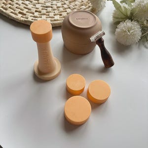 Puede incluir: Un surtido de artículos de cerámica color melocotón, que incluyen un tarro, un sello y varias piezas redondas. El sello tiene la palabra "KALIPOX" impresa. También hay un sello con mango de madera. Los artículos están dispuestos sobre una superficie blanca.