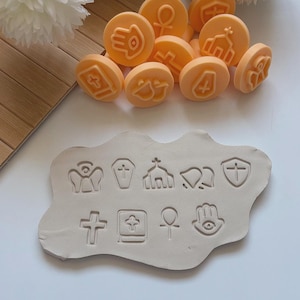 Op de afbeelding: Oranje ronde stempels met religieuze symbolen, waaronder kruisen, engelen en kerken, liggen verspreid rond een kleivorm met bijpassende afdrukken. De stempels hebben een diameter van ongeveer 2,5 cm. De kleivorm heeft verschillende religieuze symbolen erin gedrukt.