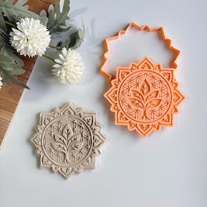 Può includere: Un tagliabiscotti arancione e un biscotto beige con un motivo floreale. Il tagliabiscotti ha la forma di un sole con una foglia dettagliata e un motivo geometrico. Il biscotto beige ha lo stesso design, mostrando l'impressione.