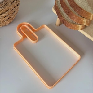 May include: An orange plastic bread-shaped cookie cutter. The cutter is in the shape of a cutting board with a handle. Slices of bread are visible in the background. The cutter is designed for making bread-shaped cookies or sandwiches.