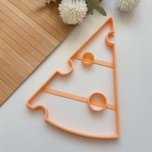 Könnte beinhalten: Ein orangefarbener Ausstecher in Form eines Käsescheibe. Er hat zwei kreisförmige Aussparungen und einen gebogenen Rand. Im Hintergrund befinden sich eine Holzoberfläche und weiße Blumen.